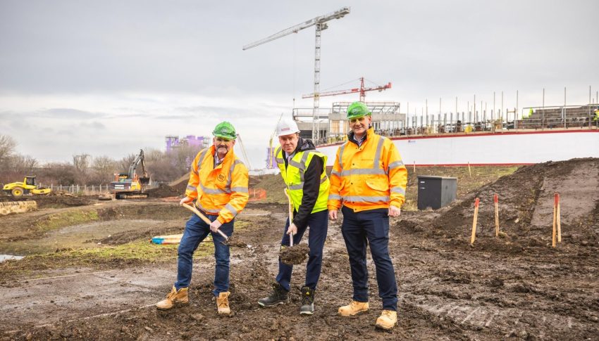 Three people stood on building site in the mud in hi vis and hard hats with shovels in their hands.