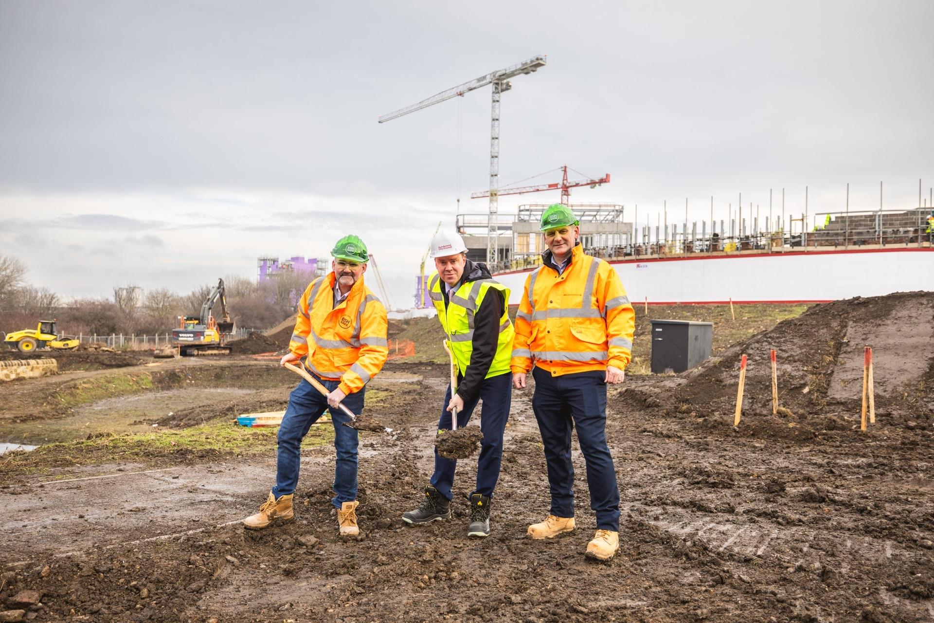 Three people stood on building site in the mud in hi vis and hard hats with shovels in their hands.